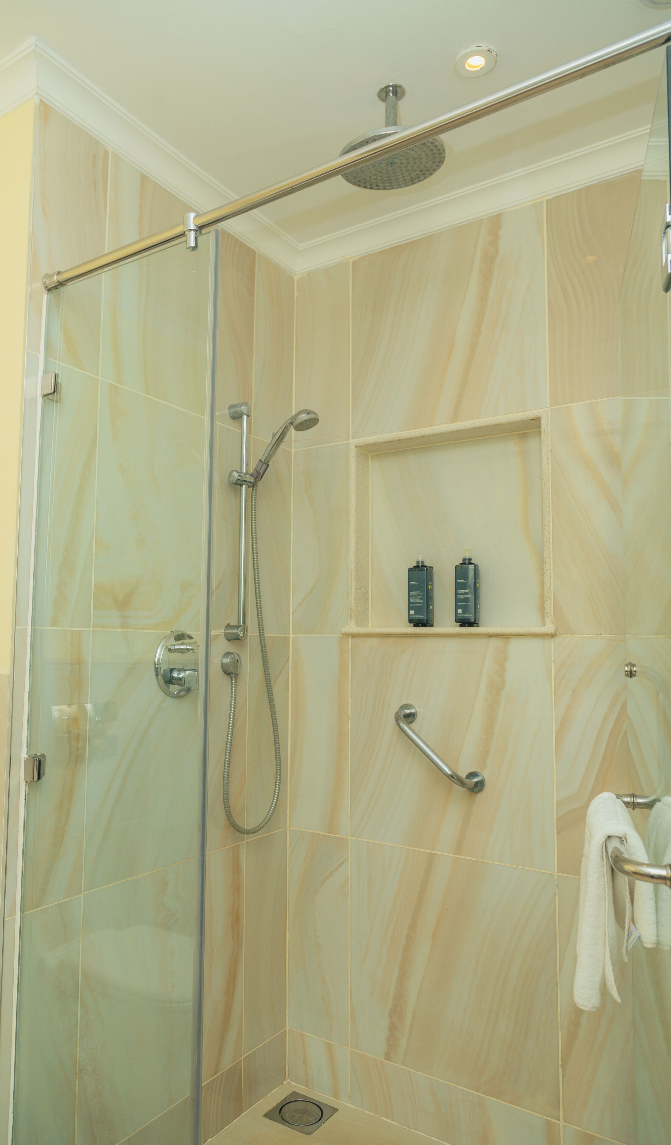A glass-enclosed shower with a handheld showerhead, a wall-mounted showerhead, and a shelf with toiletries.