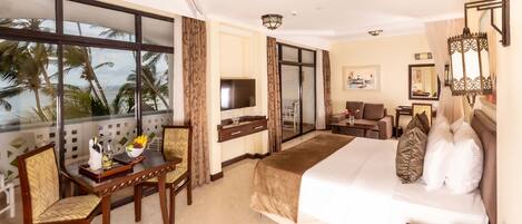 A hotel room with a bed, a desk, a chair, a TV, and a balcony with a view of palm trees and the sea.