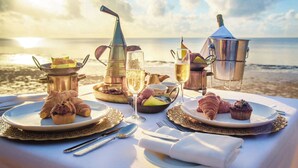 Mesa posta com croissants, muffins e taças de champanhe numa praia com vista para o pôr do sol.