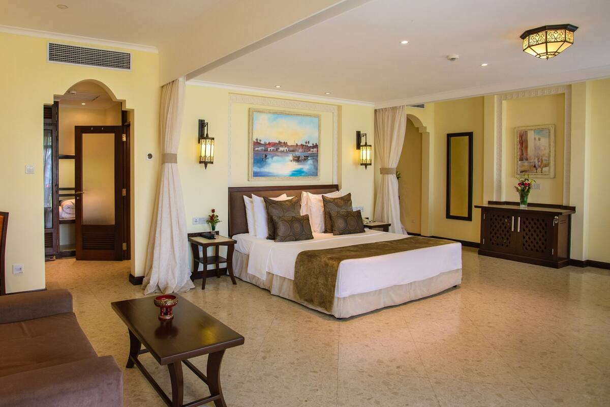 A hotel room with a large bed, a sitting area with a sofa and a table, a desk with a lamp, and a painting on the wall.