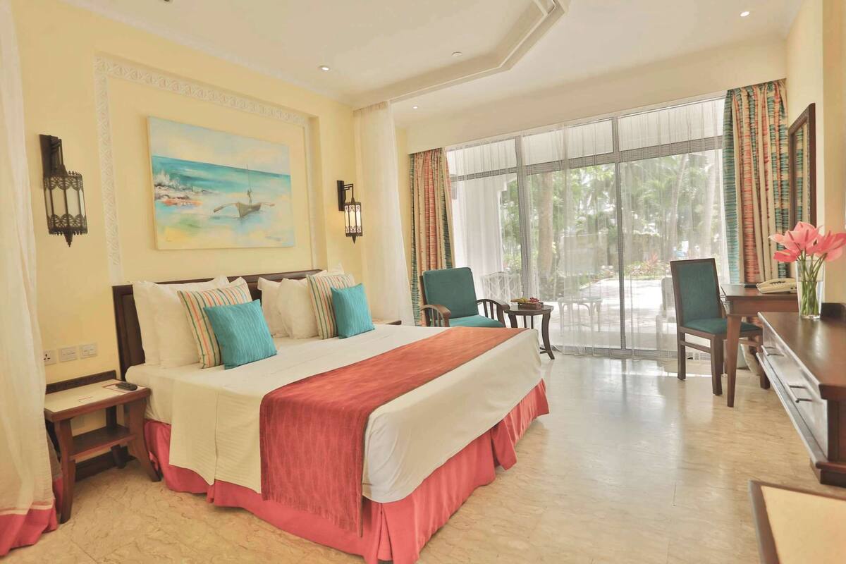 A hotel room with a large bed, a desk, a chair, a small table, and a window with curtains.