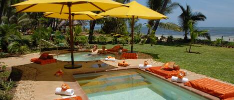 A pool area with multiple sun loungers and umbrellas, surrounded by palm trees and a view of the ocean.