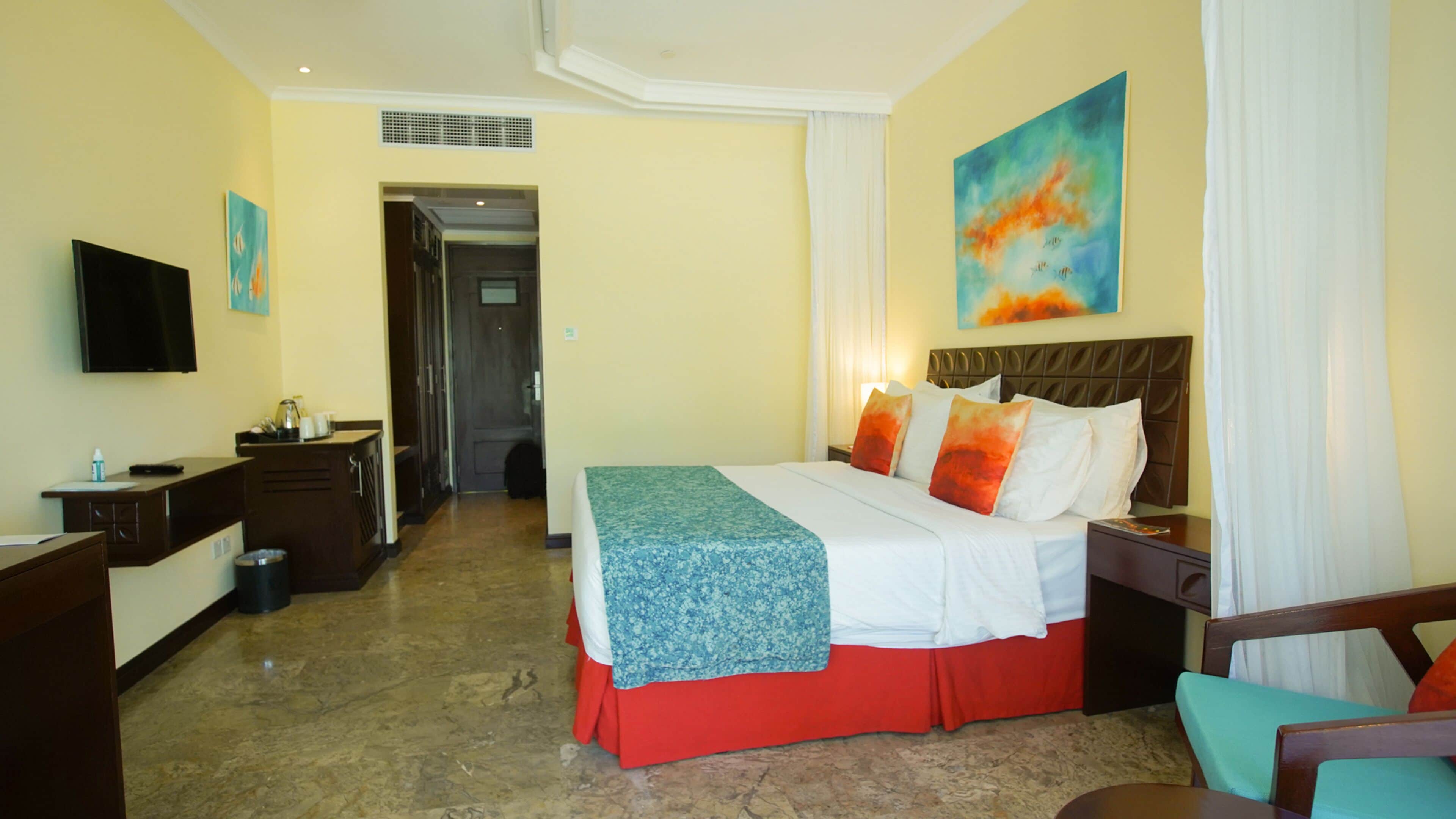 A hotel room with a bed, a desk, a TV, and a painting on the wall.