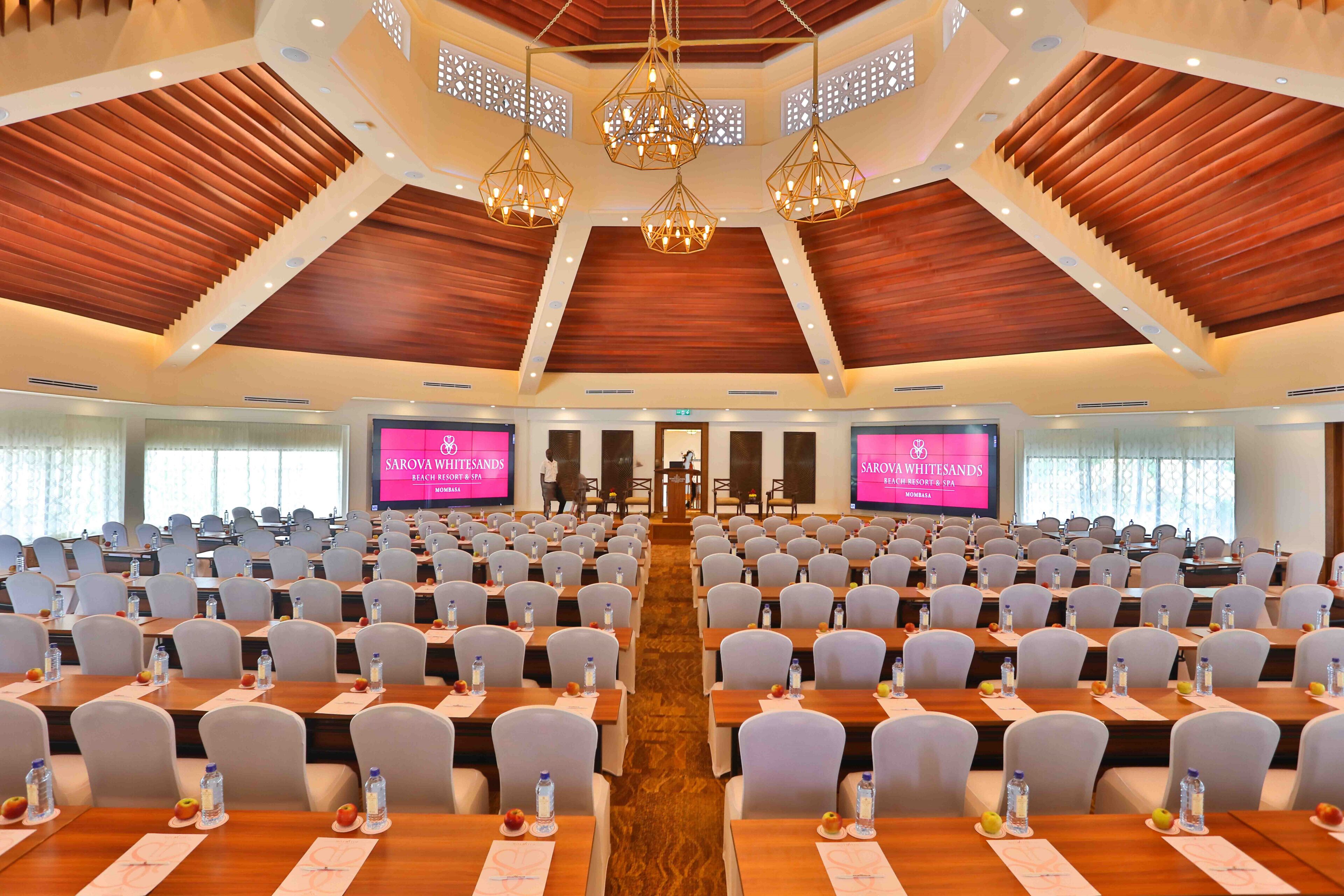 A conference room with rows of tables and chairs, a wooden ceiling, and three large screens at the front.