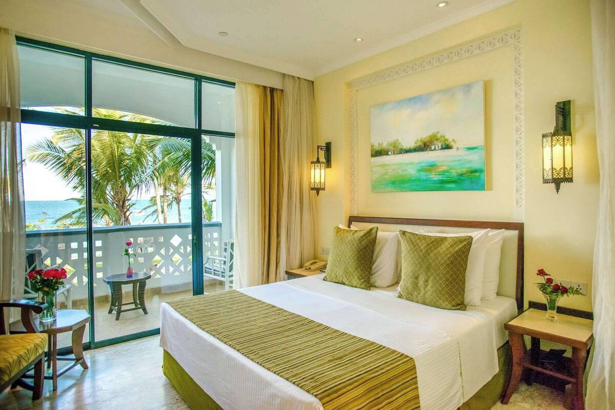 A hotel room with a large bed, a balcony with a view of the sea, and a painting on the wall.
