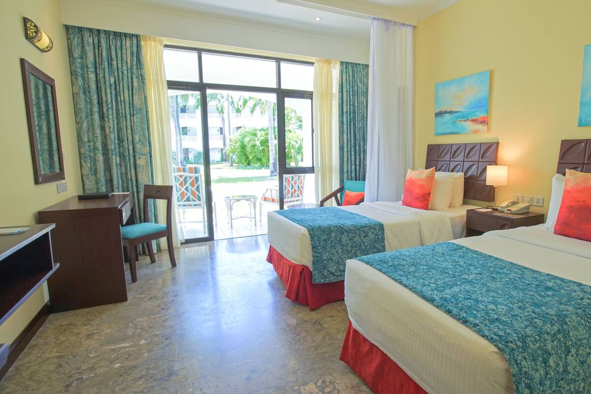 A hotel room with two beds, a desk, a chair, and a balcony with a view of palm trees.