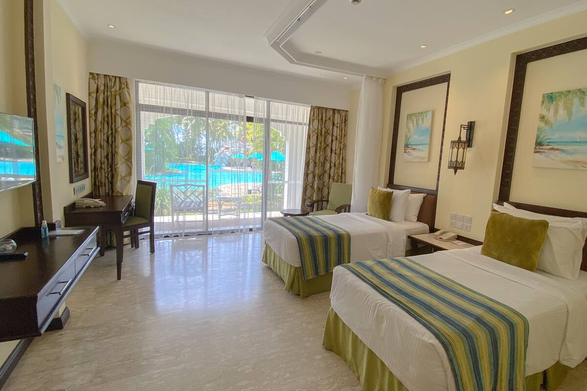 A hotel room with two beds, a desk, a TV, and a view of a pool.