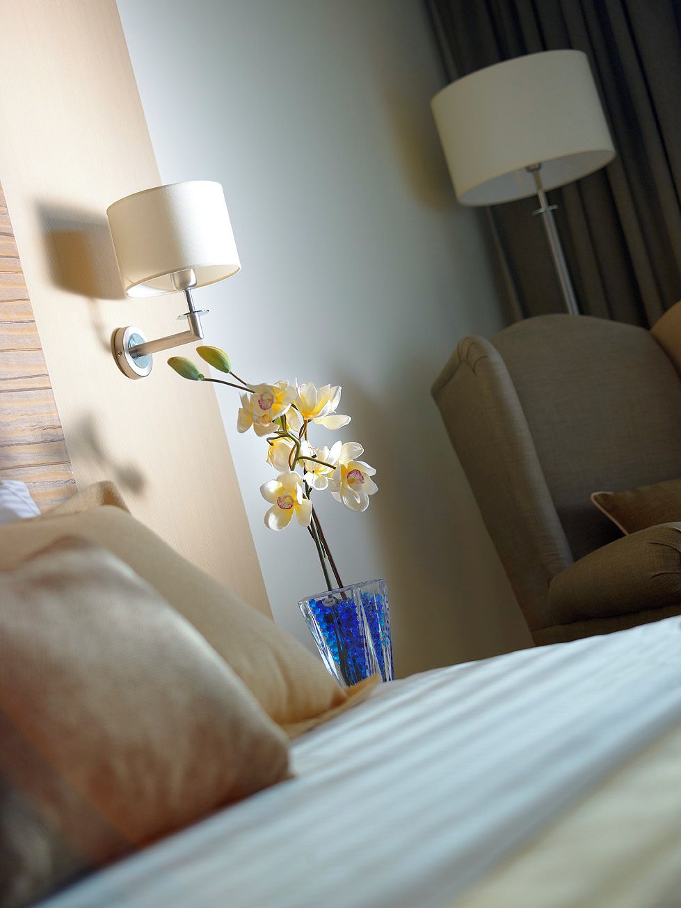 A hotel room with a bed, a nightstand with a lamp, a vase with flowers, and a chair.