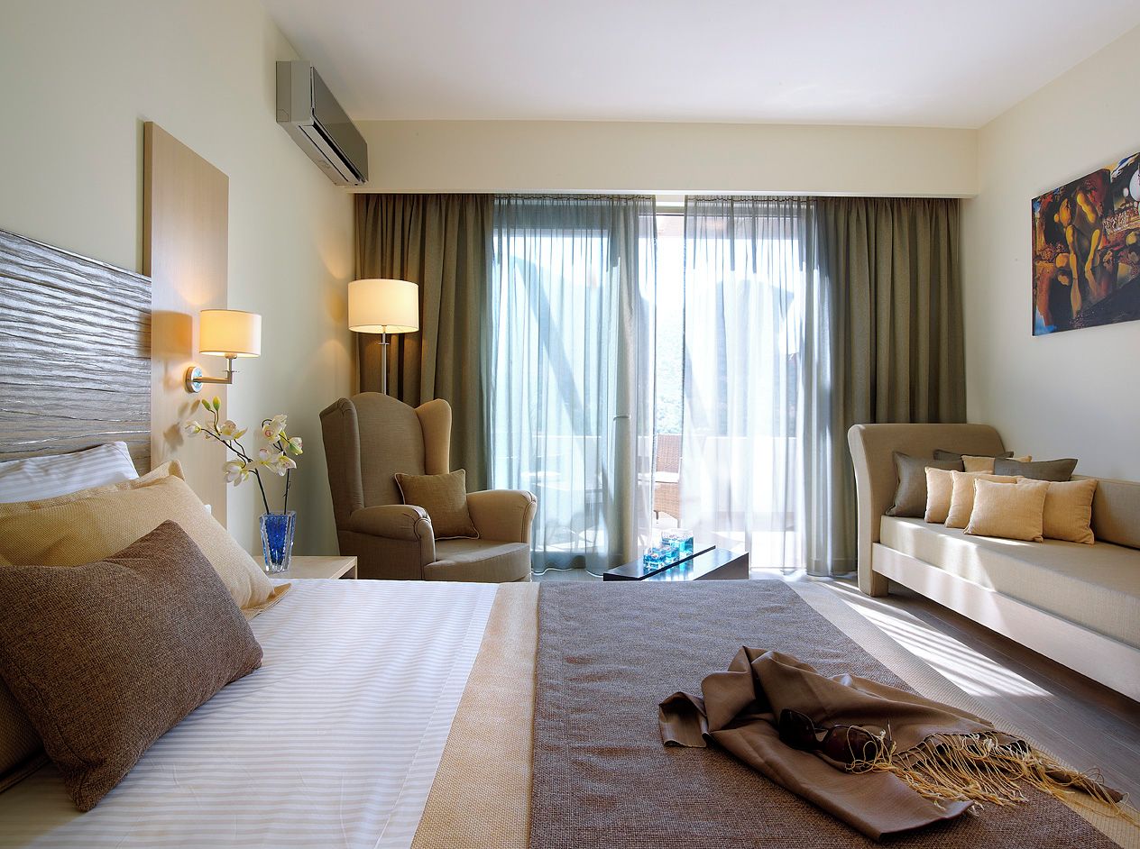Habitación de hotel con una cama grande, dos sillones, un sofá, un escritorio y un ventanal con cortinas.