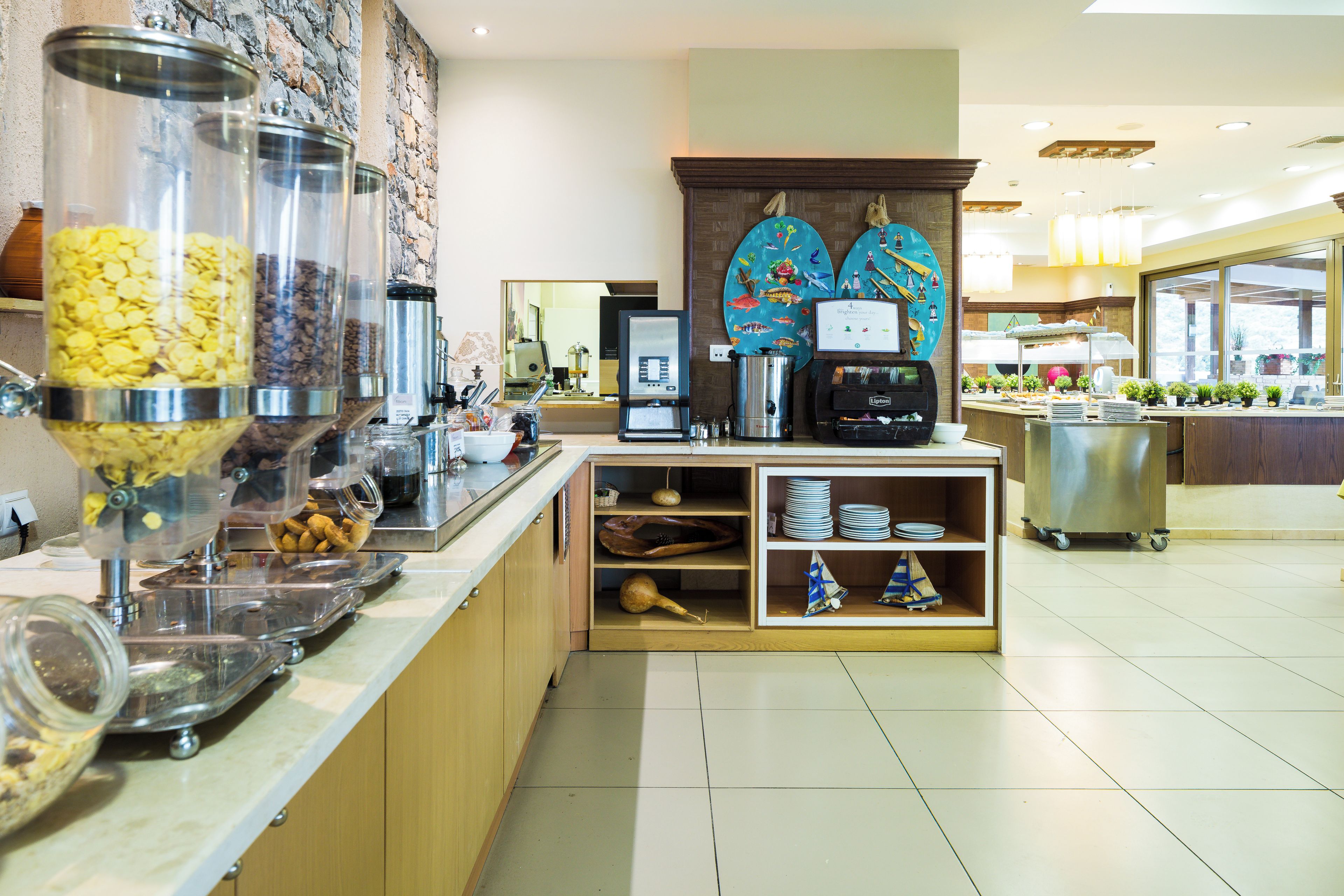 Zona de buffet con diferentes recipientes de comida, una máquina de café y una pared decorativa con temática de peces.