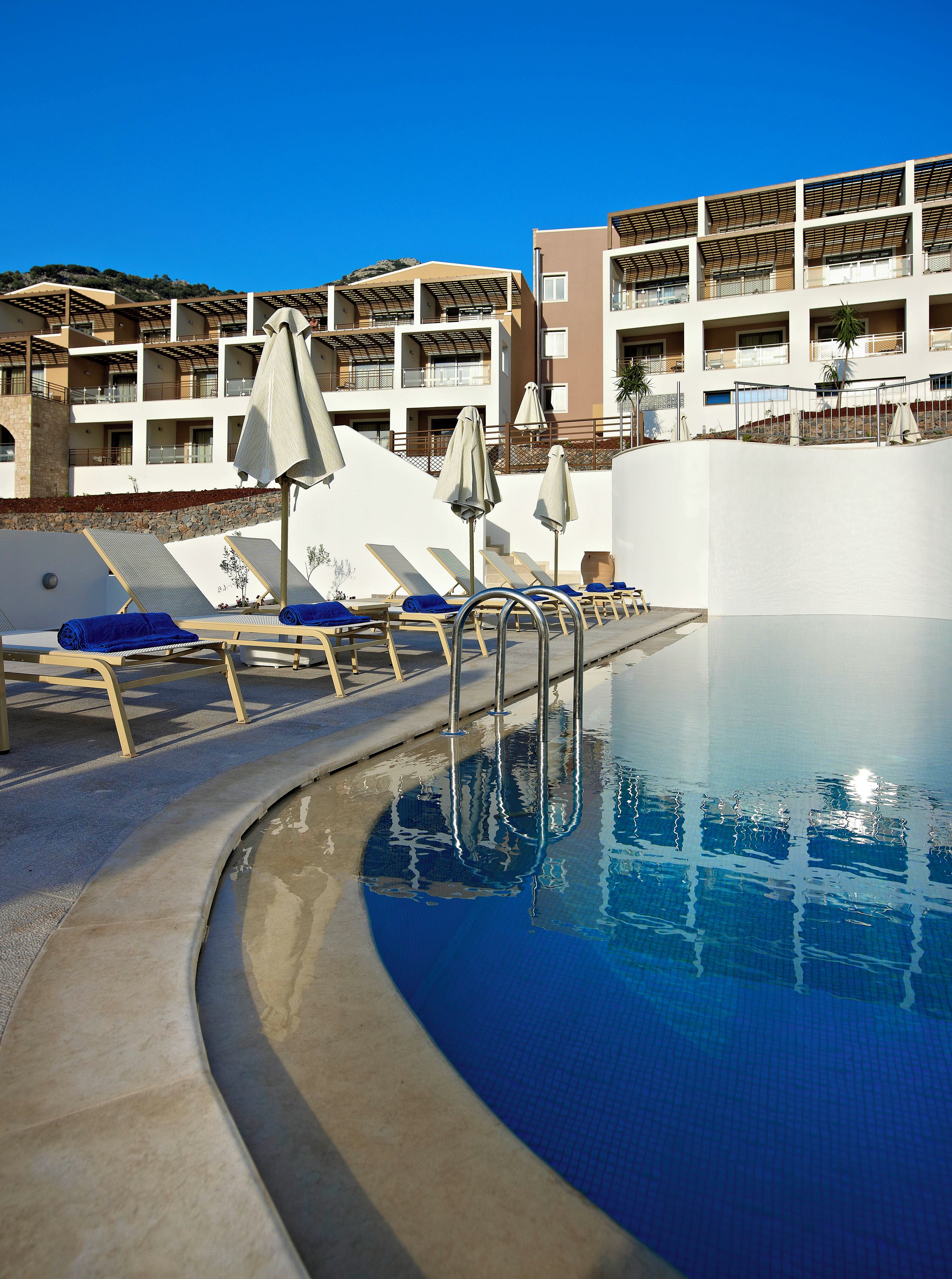 A swimming pool with sun loungers and umbrellas, surrounded by a building with balconies.