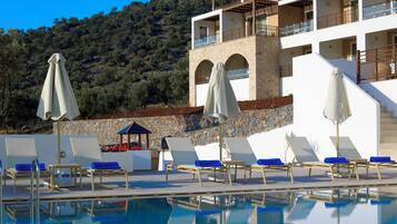 Zona de piscina con sillones reclinables y sombrillas, un muro de piedra y un edificio con balcones.