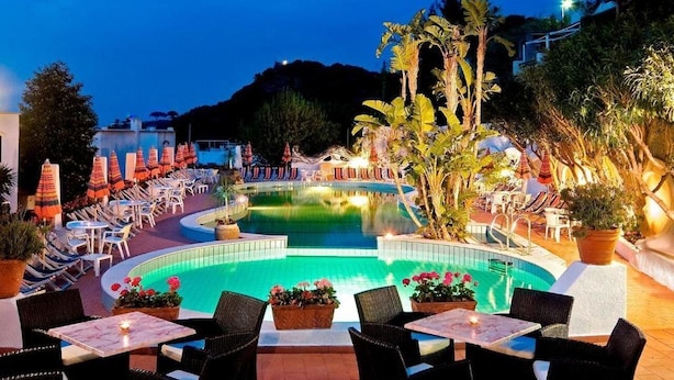 A poolside area with lounge chairs and tables, surrounded by tropical plants and a pool.