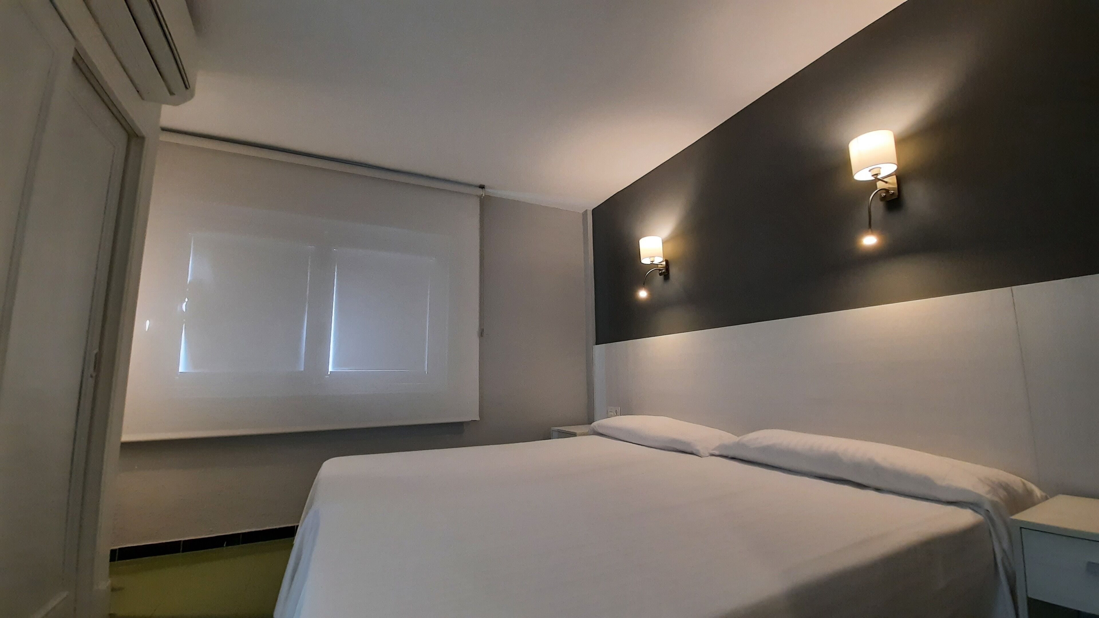 A hotel room with a bed, a nightstand, a wall-mounted lamp, and a window with blinds.