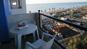 A balcony with a table and chairs, overlooking a coastal view with buildings and palm trees.