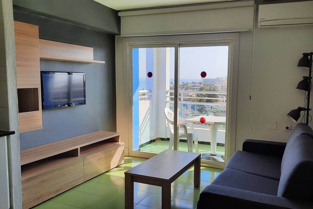A modern living room with a flat-screen TV, a sofa, a table, and a balcony with a view of the city.