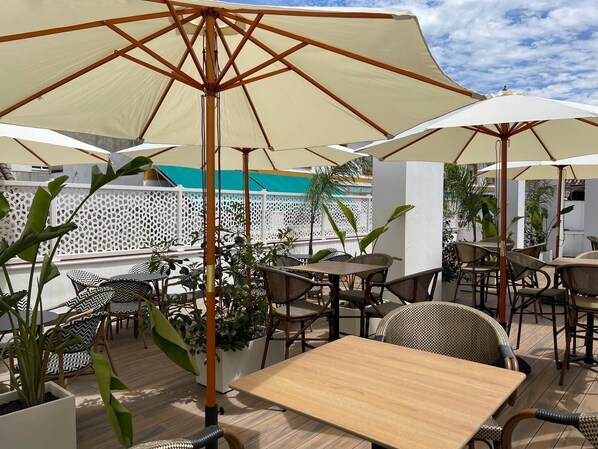 An outdoor patio with tables and chairs under large umbrellas, surrounded by potted plants and a white fence.
