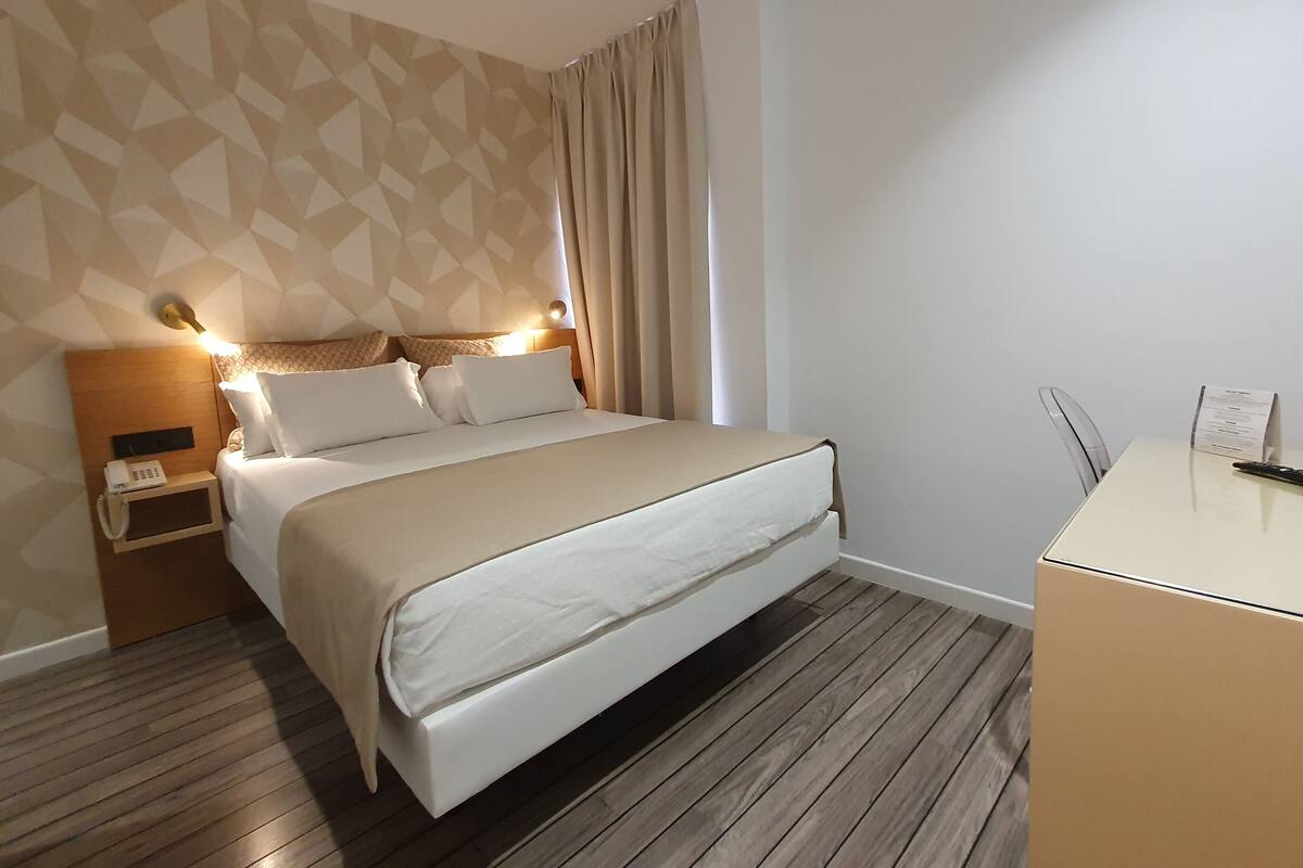 Ein Hotelzimmer mit Bett, Nachttisch, Lampe und Schreibtisch mit Stuhl.