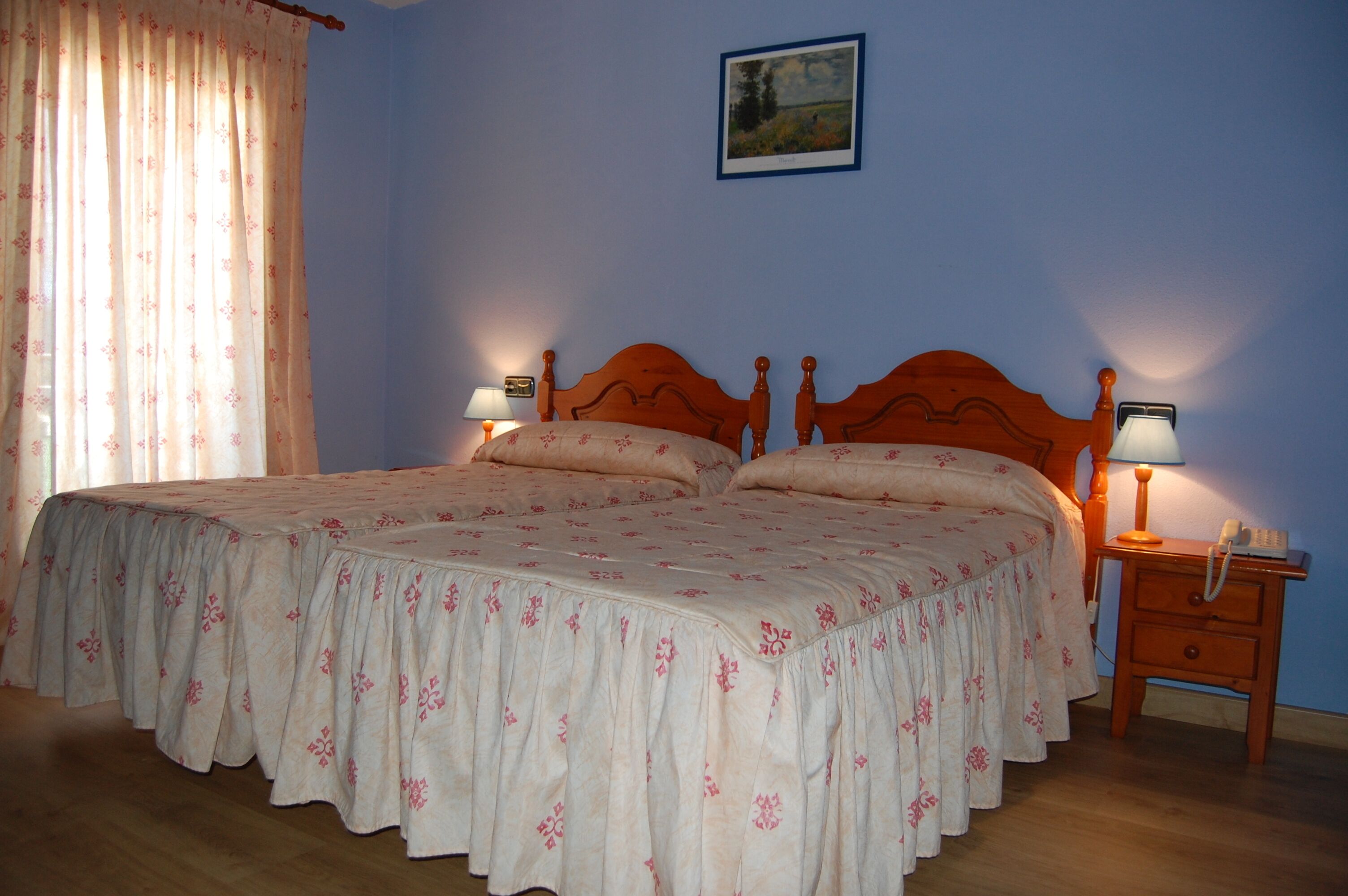 Un dormitorio con una cama doble, dos mesitas de noche con lámparas, un cabecero de madera, un cuadro enmarcado en la pared y una ventana con cortinas.
