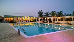 A swimming pool with a tiled deck, a covered seating area with tables and chairs, and a building with a green roof in the background. - Villa Fanusa (Syracuse)