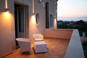 Un balcón con dos sillones blancos de mimbre y un pequeño reposapiés, una pared blanca con un ventanal circular y vistas del entorno al anochecer.