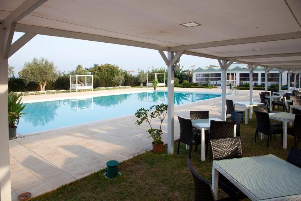 A covered outdoor pool area with lounge chairs and tables, surrounded by greenery and a building in the background.