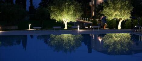 A nighttime view of a swimming pool with trees and a building in the background.