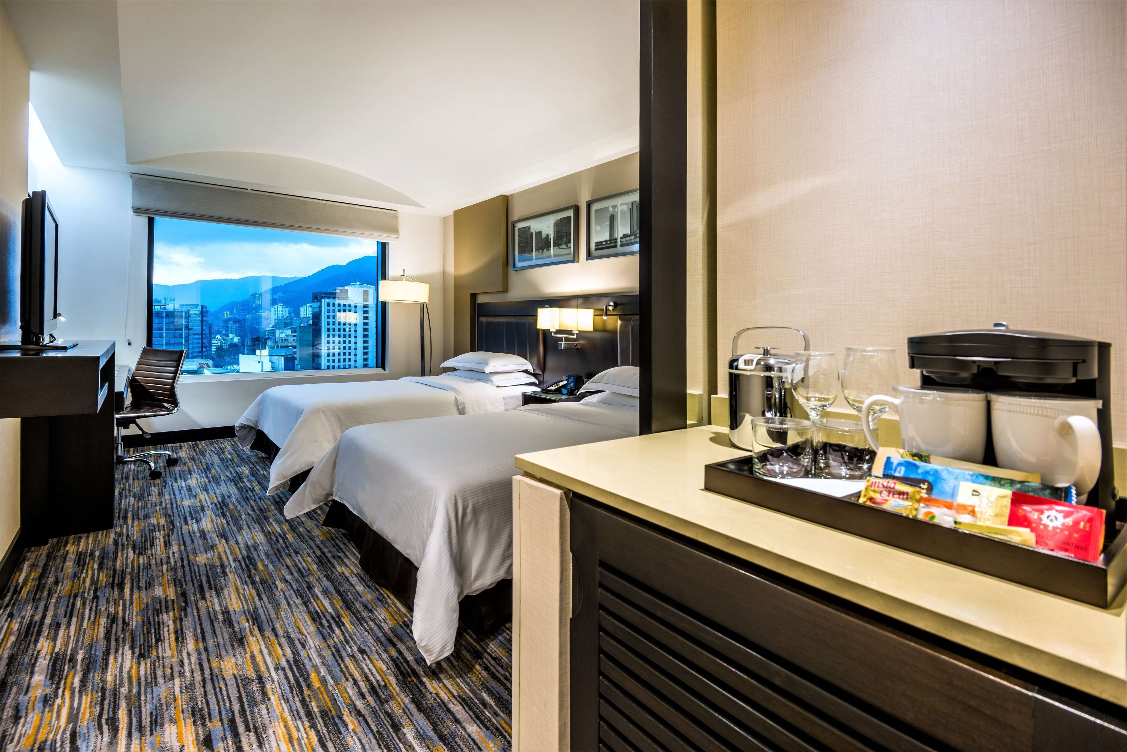 Habitación de hotel con dos camas, un escritorio con cafetera, un televisor y un ventanal con vistas a la ciudad.