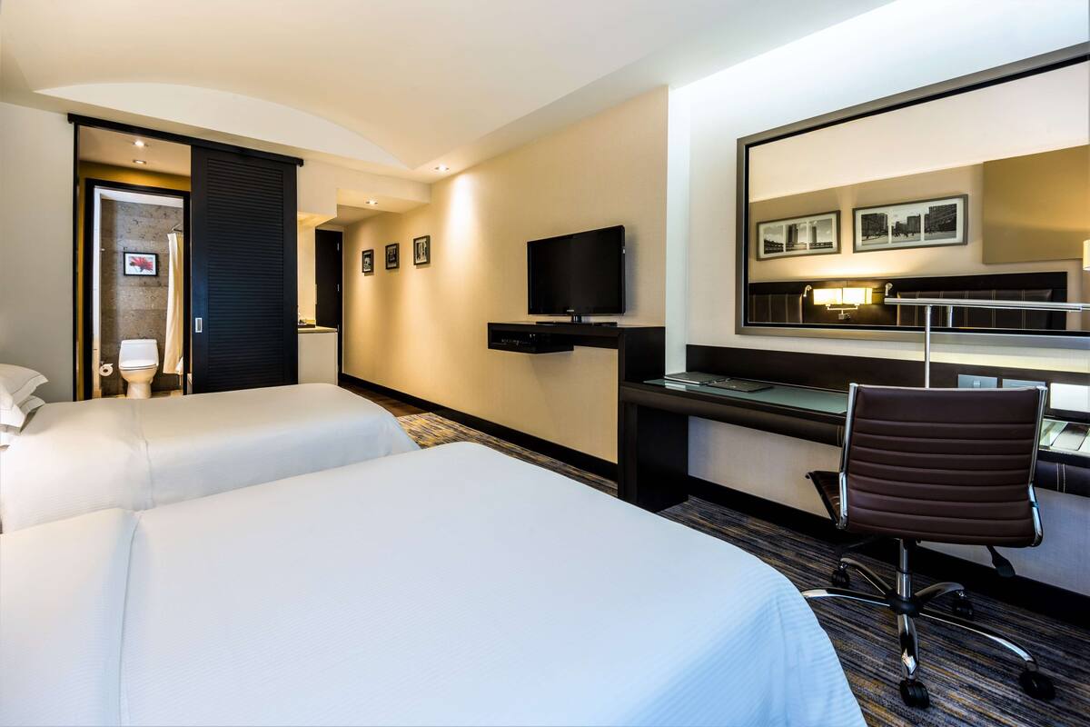 A hotel room with two beds, a desk with a flat-screen TV, a chair, and a large mirror.