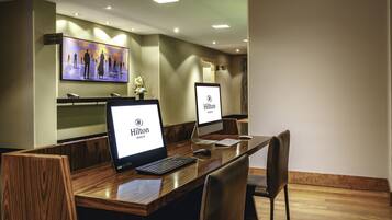 A hotel reception desk with two computer monitors displaying the Hilton logo, a wooden reception desk, and a television screen in the background.