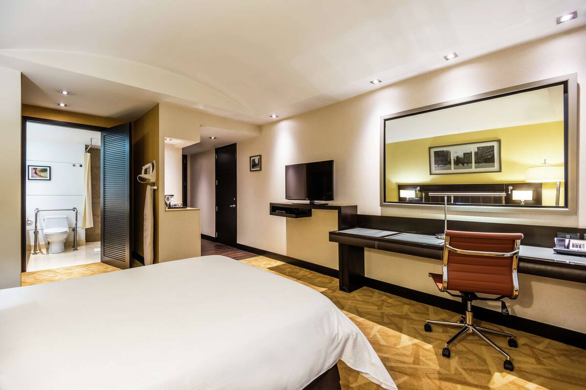 A hotel room with a large bed, a desk with a chair, a television, and a bathroom visible through an open door.