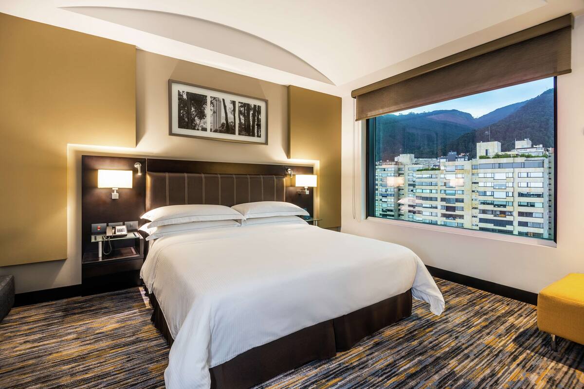 A hotel room with a large bed, a nightstand, a desk, and a window with a city view.