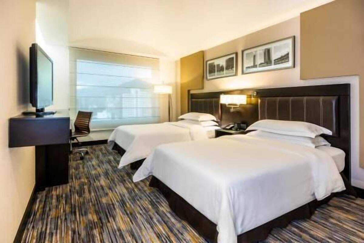A hotel room with two beds, a television, a desk, and a window with a view.