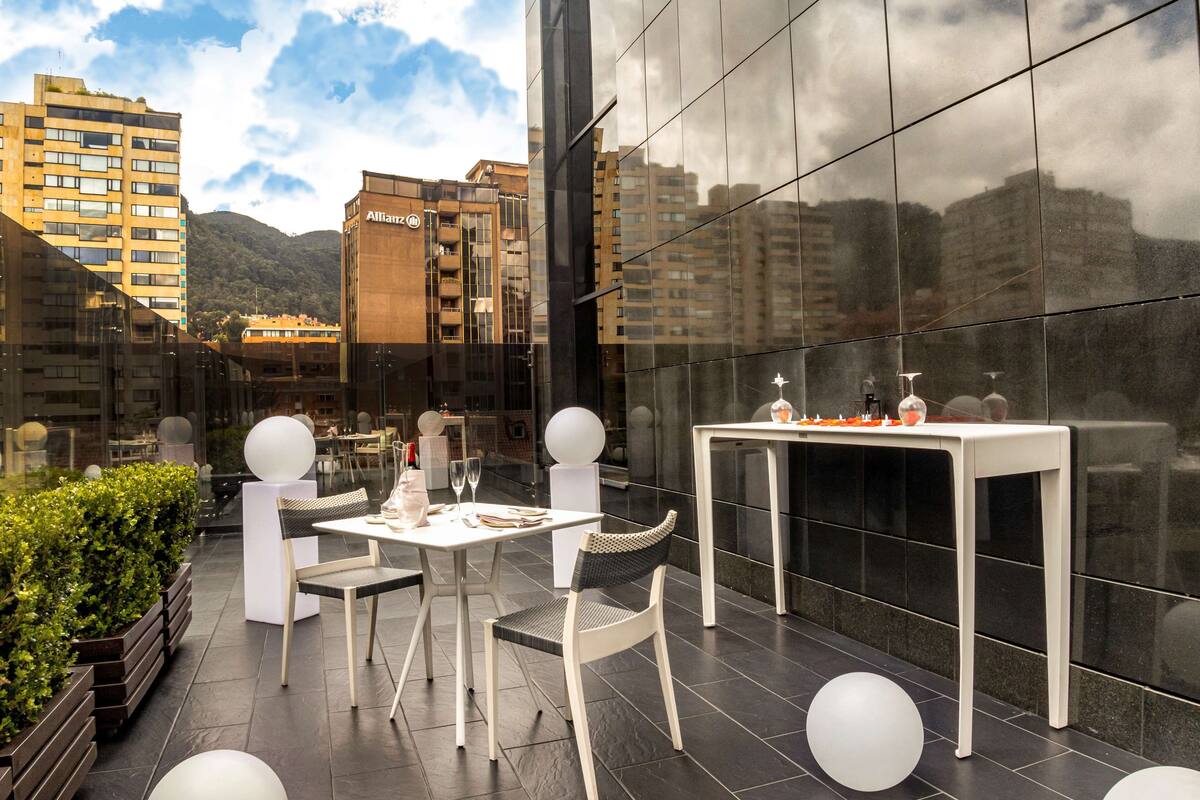 An outdoor dining area with a table and chairs, a small white table with bottles, and a view of buildings and mountains in the background.