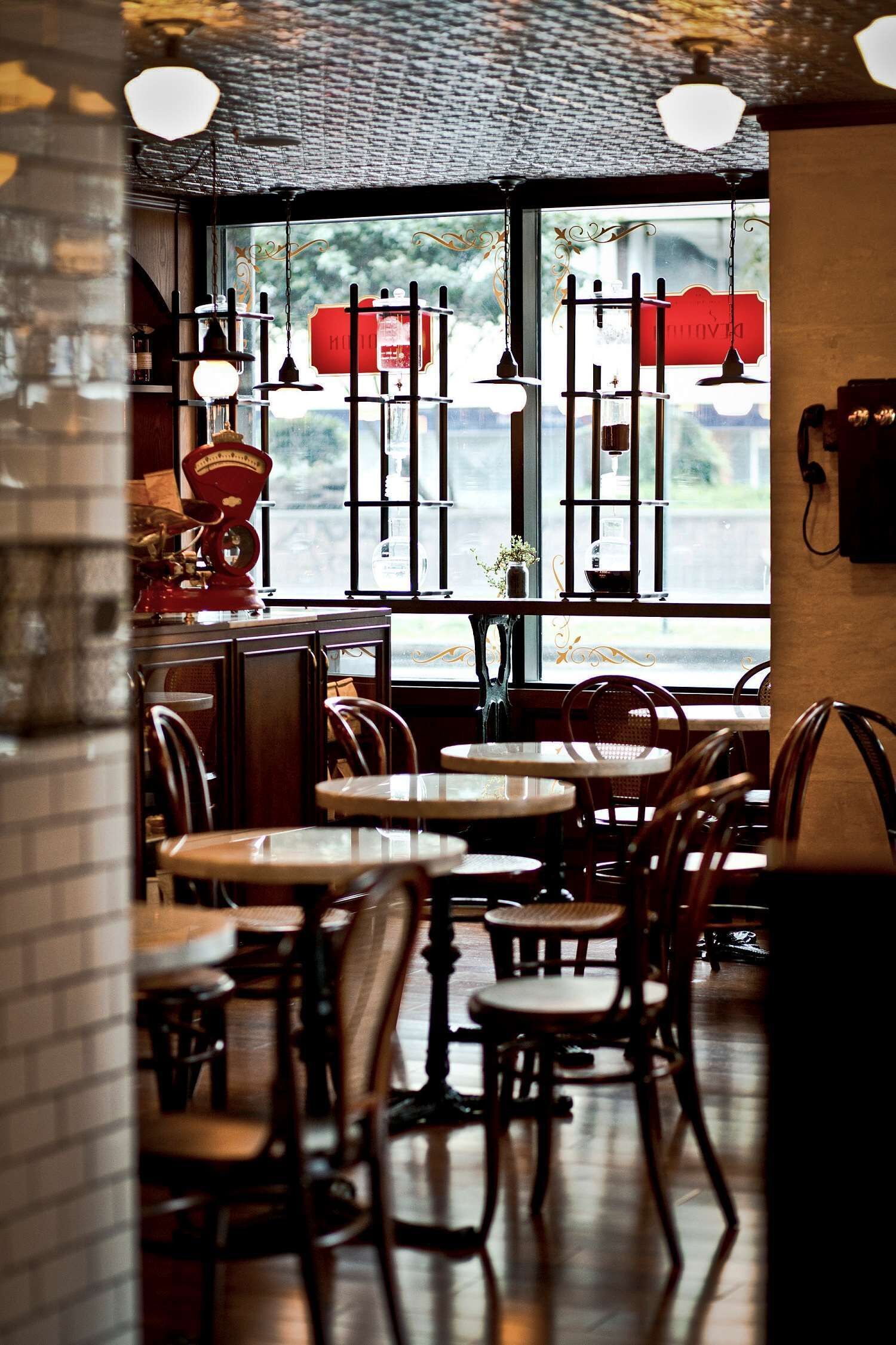 A cozy cafe interior with wooden tables and chairs, hanging light fixtures, and a view through a window with a red sign.