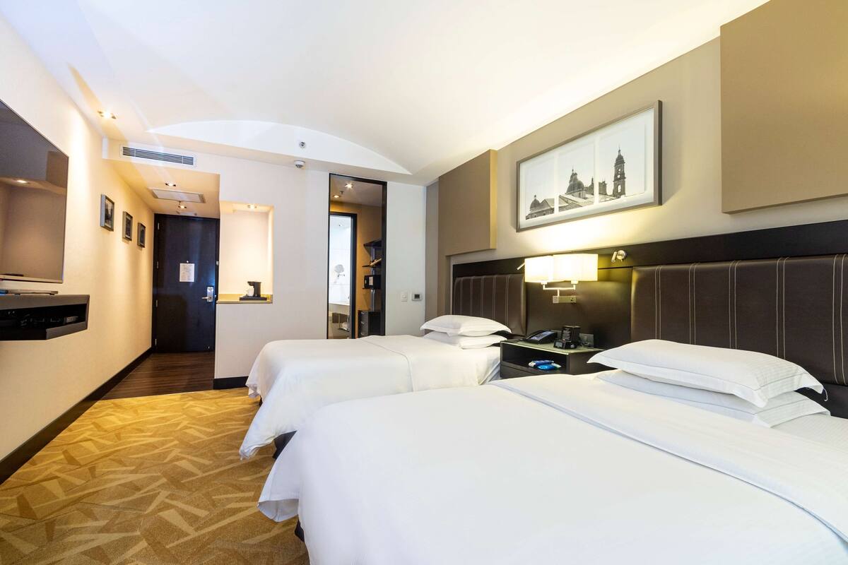A hotel room with two beds, a desk with a lamp, a picture on the wall, and a hallway leading to another room.