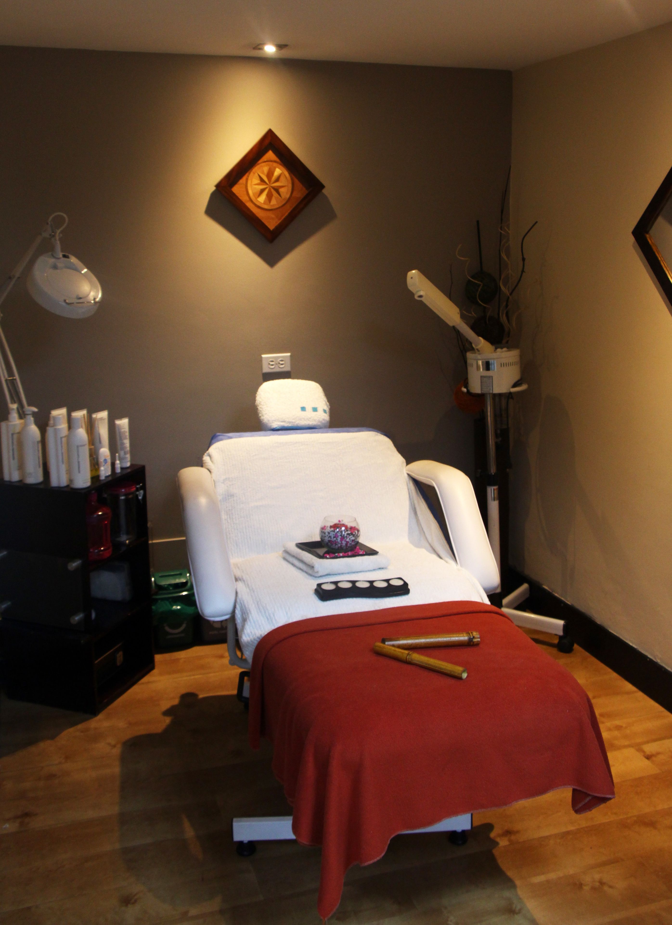 A massage therapy room with a massage table, a red blanket, a foot massager, a small decorative bowl, and a shelf with bottles.