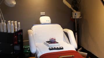 A massage therapy room with a massage table, a red blanket, a foot massager, a small decorative bowl, and a shelf with bottles.