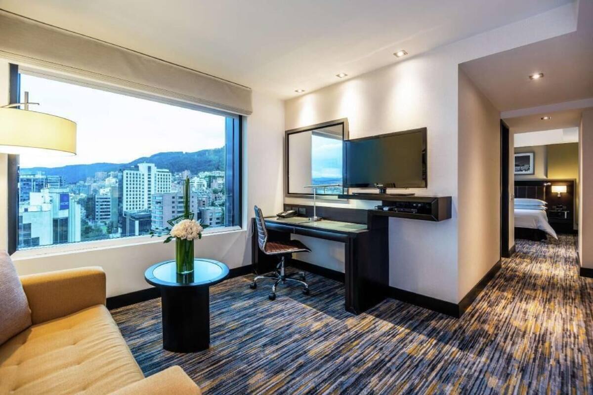 A hotel room with a large window, a desk, a chair, a sofa, and a television.
