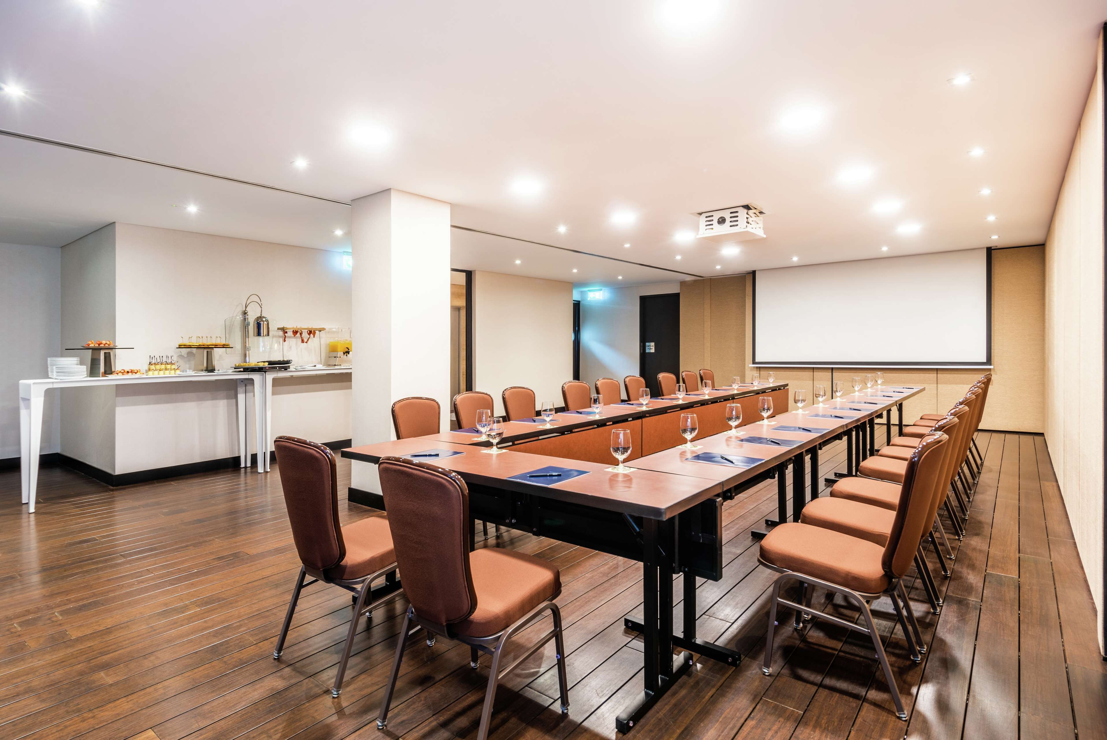Une salle de conférence équipée d’une longue table, de chaises, d’un écran de projection et d’un espace buffet.