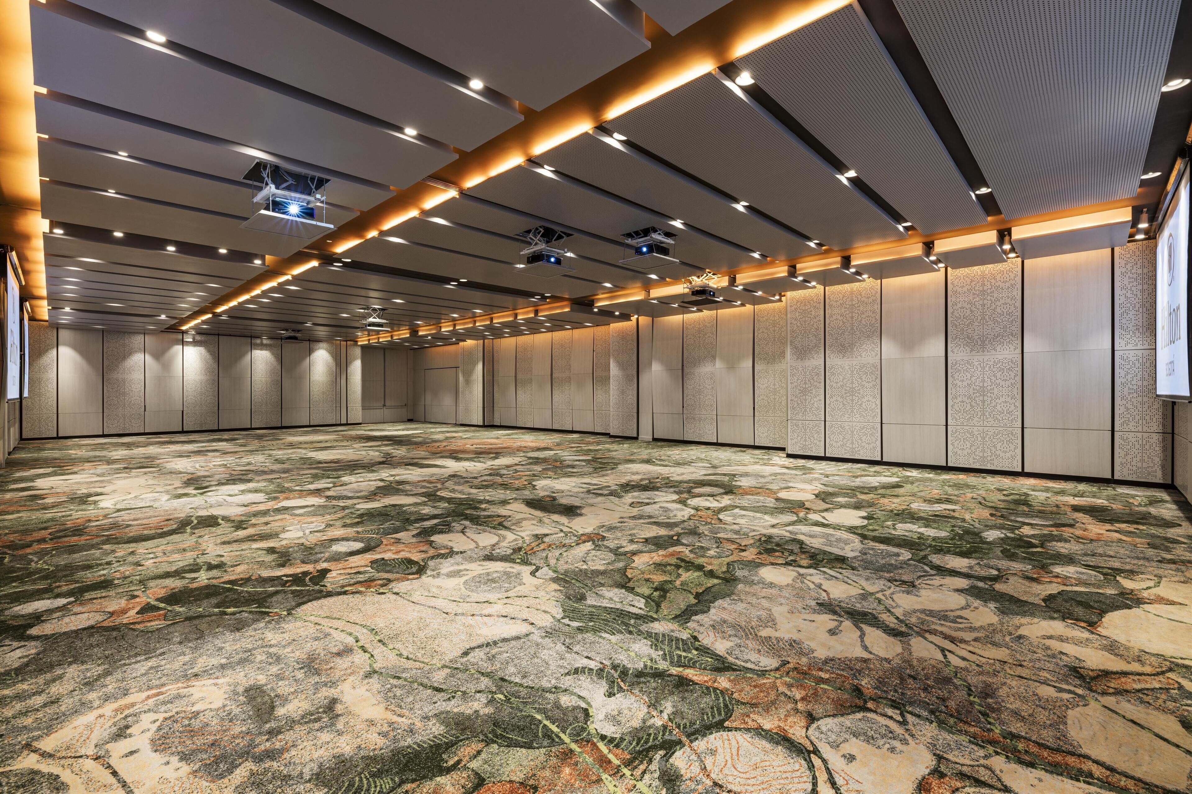 Una amplia sala de conferencias con alfombra estampada, iluminación empotrada y un alto techo con una rejilla de luces y proyectores.