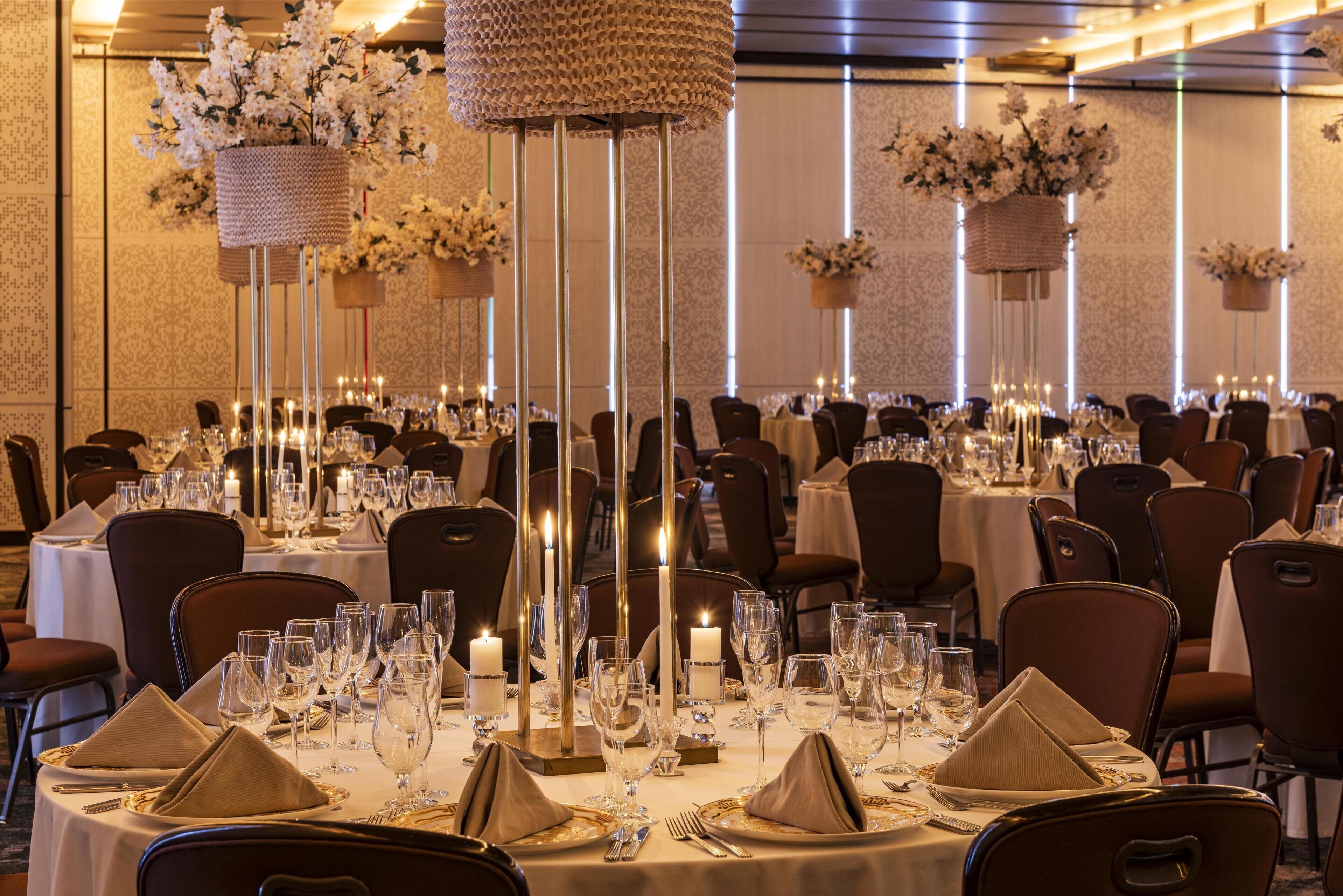 A banquet hall with round tables set for dining, featuring white tablecloths, gold napkins, and tall candle holders.