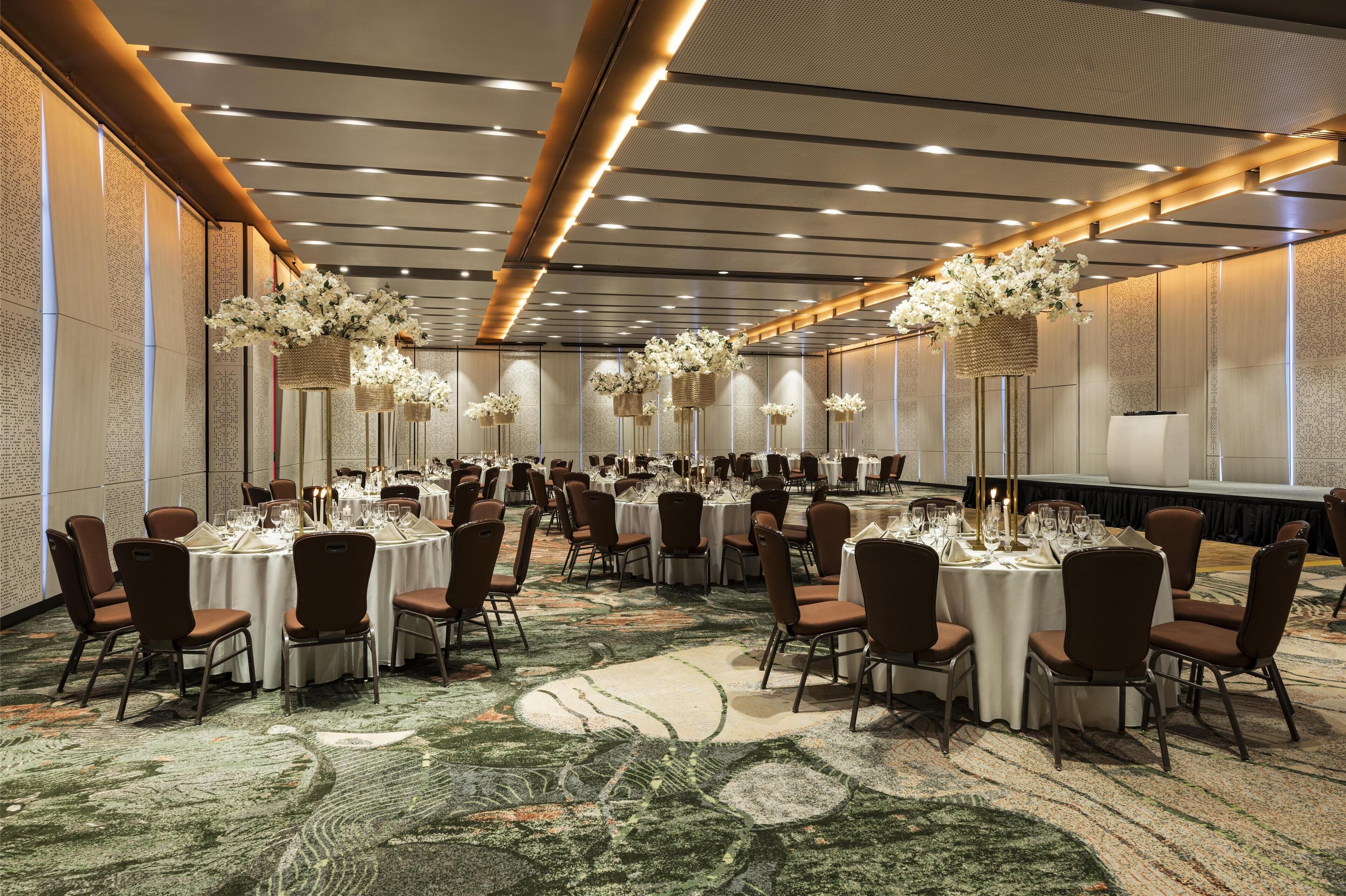 A banquet hall with round tables set for dining, featuring a carpeted floor and floral centerpieces.