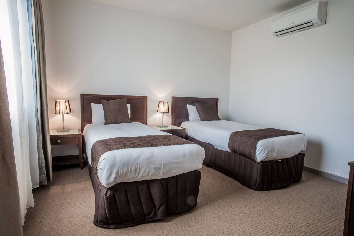 A hotel room with two beds, each with a wooden headboard, a nightstand, a lamp, and an air conditioning unit on the wall.