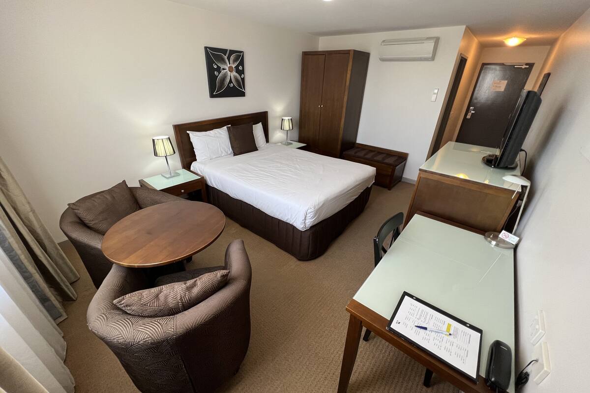 A hotel room with a bed, a sofa, a desk, and a chair. There is a nightstand with a lamp, a wardrobe, and a TV.