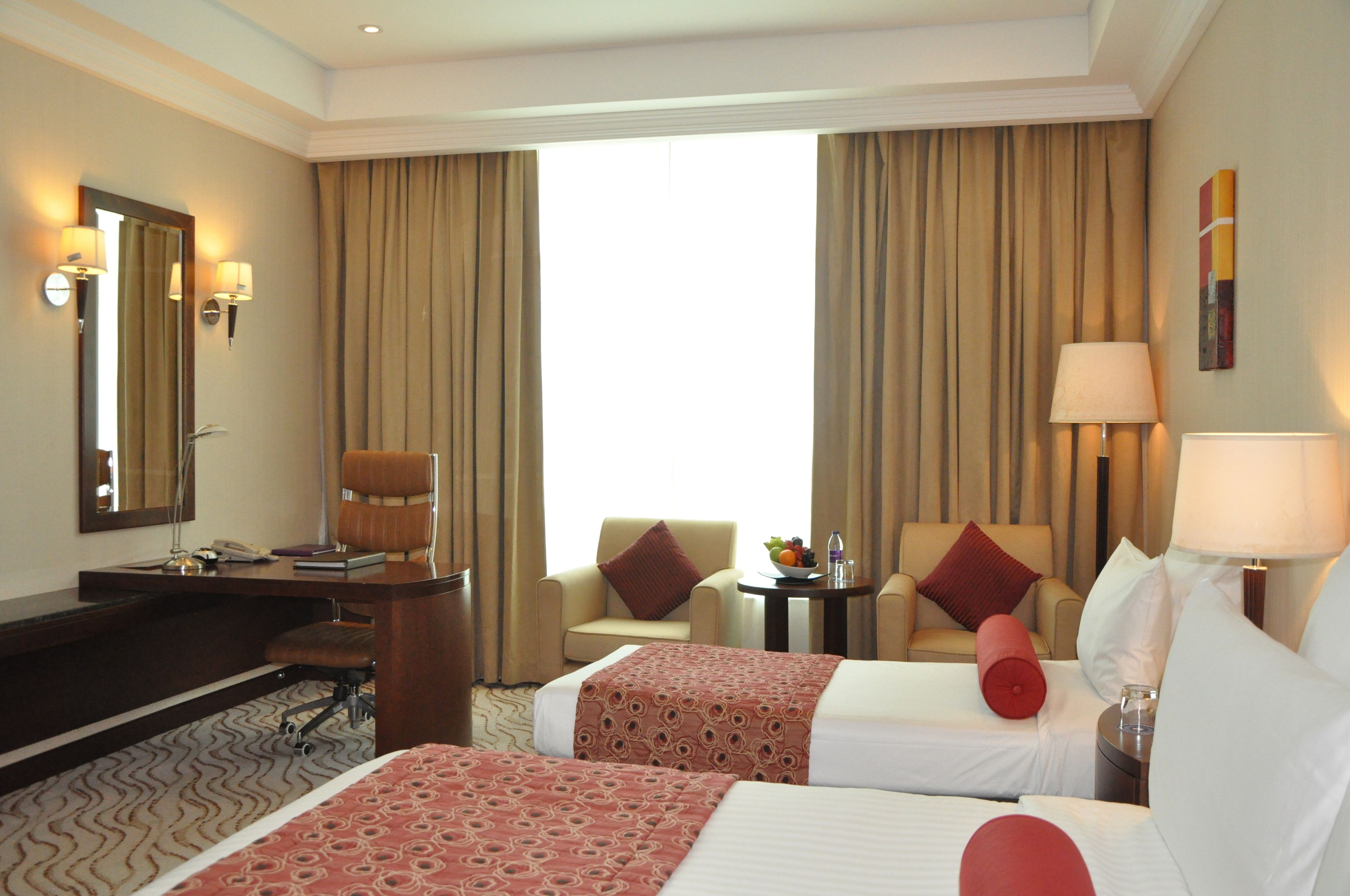 A hotel room with two beds, a desk, a chair, a small table with a lamp, and a large window with curtains.