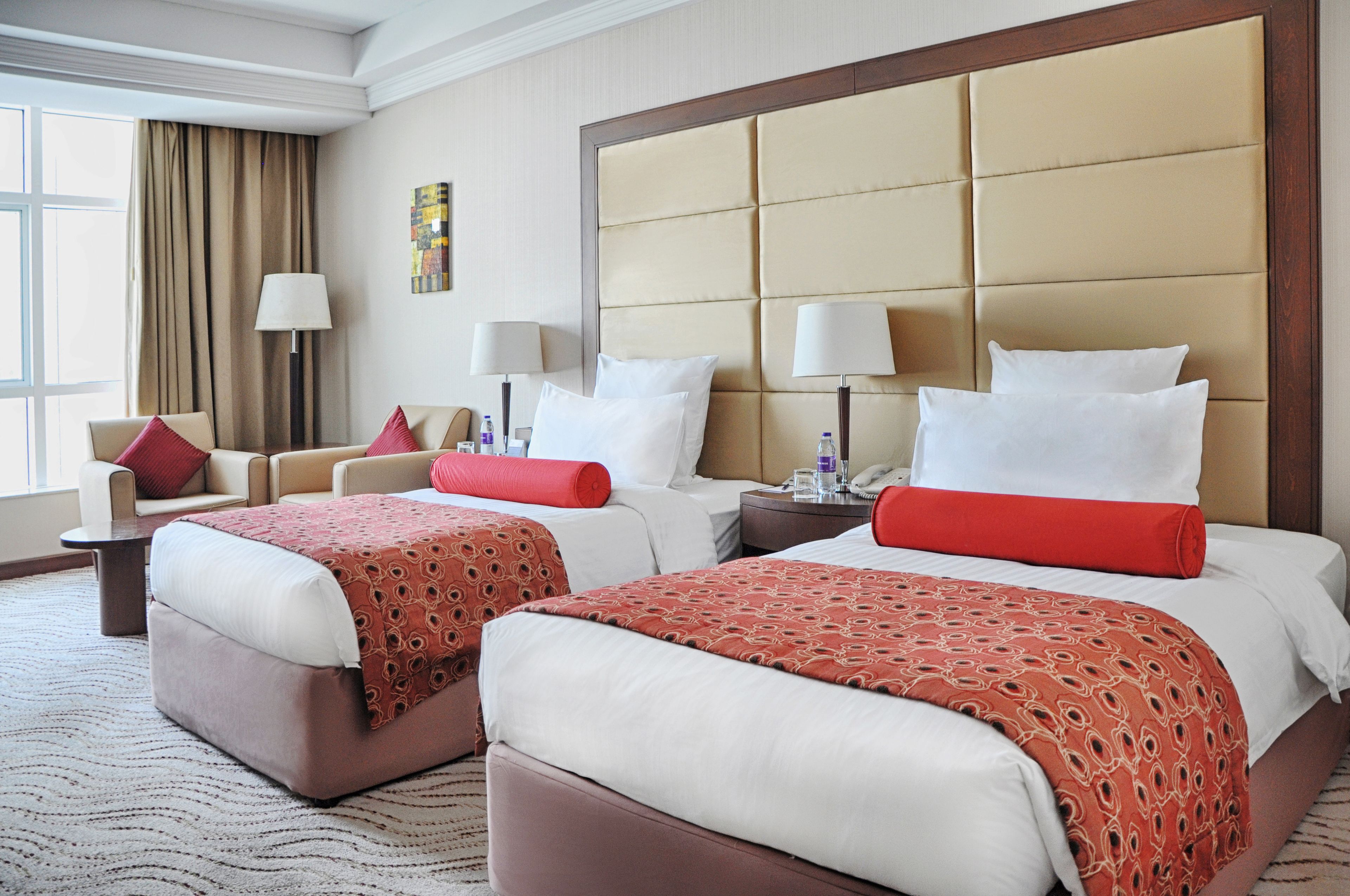 A hotel room with two beds, a sofa, a nightstand with a lamp, and a view of the outside.