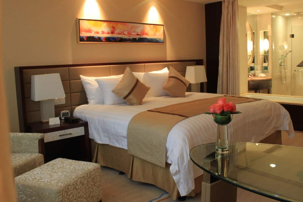 Une chambre d’hôtel avec un lit, des lampes de chevet, une table en verre ornée d’un vase de fleurs, une table de chevet, une chaise et un grand miroir.