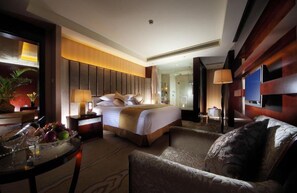 Ein Hotelzimmer mit Bett, Sitzgelegenheit mit Stuhl, einem kleinen Tisch mit Obst und Gläsern sowie einem Fernseher.