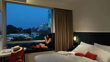 Habitación de hotel con un ventanal grande, cama con almohadas blancas y rojas, una silla roja y una mesita con un sombrero y un libro.